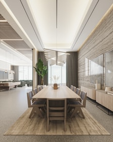 A modern dining room features a long wooden table with eight chairs placed around it. The room is well-lit with a unique, geometric ceiling light fixture. A large plant is situated near the floor-to-ceiling windows, which are draped with heavy curtains. On one side, there is a sideboard adorned with decorative art pieces and a textured wall with framed art. The space extends into a sitting area with a pool table in the background.