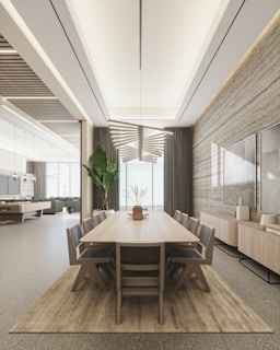 A modern dining room features a long wooden table with eight chairs placed around it. The room is well-lit with a unique, geometric ceiling light fixture. A large plant is situated near the floor-to-ceiling windows, which are draped with heavy curtains. On one side, there is a sideboard adorned with decorative art pieces and a textured wall with framed art. The space extends into a sitting area with a pool table in the background.