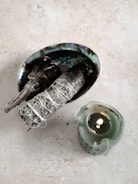 A small oval dish, intricately designed with iridescent colors, contains two smudging bundles tied with white string. Nearby, a green candle is lit, providing a small, warm flame.