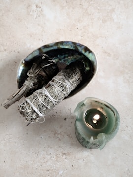 A small oval dish, intricately designed with iridescent colors, contains two smudging bundles tied with white string. Nearby, a green candle is lit, providing a small, warm flame.