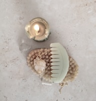A lit candle in a greenish holder sits beside a translucent stone on a light surface. Below them is a natural bristle brush and a large gemstone necklace, with a pastel green comb placed on top, all arranged on a stone-textured background.