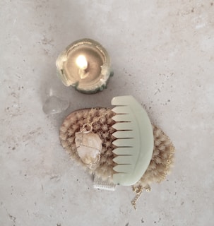 A lit candle in a greenish holder sits beside a translucent stone on a light surface. Below them is a natural bristle brush and a large gemstone necklace, with a pastel green comb placed on top, all arranged on a stone-textured background.