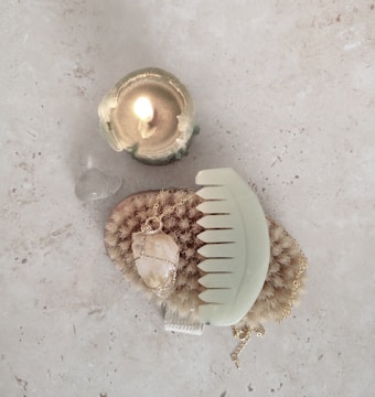 A lit candle in a greenish holder sits beside a translucent stone on a light surface. Below them is a natural bristle brush and a large gemstone necklace, with a pastel green comb placed on top, all arranged on a stone-textured background.