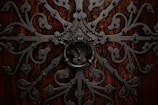 Intricate wrought iron design with elaborate scrollwork and a large circular handle set against a rich, dark wooden background. The symmetrical pattern features floral and geometric elements, creating an ornate and elaborate texture.