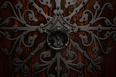 Intricate wrought iron design with elaborate scrollwork and a large circular handle set against a rich, dark wooden background. The symmetrical pattern features floral and geometric elements, creating an ornate and elaborate texture.