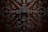 Intricate wrought iron design with elaborate scrollwork and a large circular handle set against a rich, dark wooden background. The symmetrical pattern features floral and geometric elements, creating an ornate and elaborate texture.