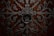 Intricate wrought iron design with elaborate scrollwork and a large circular handle set against a rich, dark wooden background. The symmetrical pattern features floral and geometric elements, creating an ornate and elaborate texture.