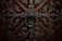 Intricate wrought iron design with elaborate scrollwork and a large circular handle set against a rich, dark wooden background. The symmetrical pattern features floral and geometric elements, creating an ornate and elaborate texture.