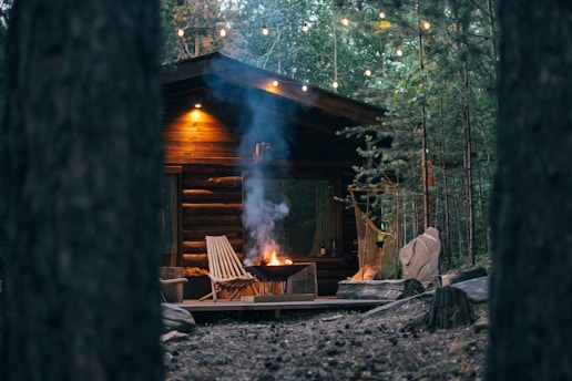 A cozy wooden cabin in a forest setting, illuminated by warm lights. It features a deck with a fire pit emanating smoke and a couple of chairs. Surrounded by lush green trees, the environment is serene and inviting.