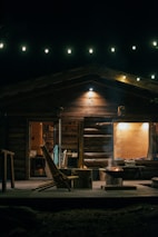 A cozy wooden cabin is illuminated by string lights at night. The front porch features wooden chairs and a small fire pit emitting gentle smoke. The interior is warmly lit, revealing bookshelves and a dining area. The setting conveys a sense of warmth and tranquility.