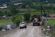 A rural scene with a dirt road where a car is parked on one side and a tractor with hay bales is being driven down the road. Several people are interacting near the car, and pigs are wandering on the roadside. The background features lush greenery, farmhouses, and some distant hills.