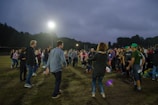 A group of people are gathered outdoors on a field, possibly engaging in a social or community event. Many individuals are dressed casually in jeans and jackets, and they appear to be conversing or participating in an activity together. The sky is overcast, and there are bright floodlights illuminating the area. Trees line the horizon in the background.