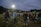 A group of people are gathered outdoors on a field, possibly engaging in a social or community event. Many individuals are dressed casually in jeans and jackets, and they appear to be conversing or participating in an activity together. The sky is overcast, and there are bright floodlights illuminating the area. Trees line the horizon in the background.