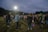 A group of people are gathered outdoors on a field, possibly engaging in a social or community event. Many individuals are dressed casually in jeans and jackets, and they appear to be conversing or participating in an activity together. The sky is overcast, and there are bright floodlights illuminating the area. Trees line the horizon in the background.
