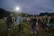 A group of people are gathered outdoors on a field, possibly engaging in a social or community event. Many individuals are dressed casually in jeans and jackets, and they appear to be conversing or participating in an activity together. The sky is overcast, and there are bright floodlights illuminating the area. Trees line the horizon in the background.