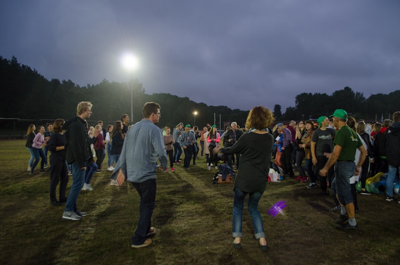 A group of people are gathered outdoors on a field, possibly engaging in a social or community event. Many individuals are dressed casually in jeans and jackets, and they appear to be conversing or participating in an activity together. The sky is overcast, and there are bright floodlights illuminating the area. Trees line the horizon in the background.