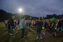 A group of people are gathered outdoors on a field, possibly engaging in a social or community event. Many individuals are dressed casually in jeans and jackets, and they appear to be conversing or participating in an activity together. The sky is overcast, and there are bright floodlights illuminating the area. Trees line the horizon in the background.
