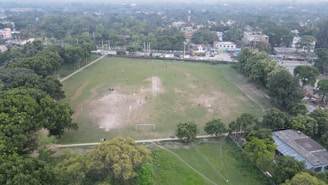 An aerial view of a large open field surrounded by trees and urban structures. The field has visible patches of grass and dirt, with people scattered across, involved in various activities. A couple of goalposts suggest the area may be used for sports. Residential and commercial buildings are visible in the background, with a road running parallel to the field.