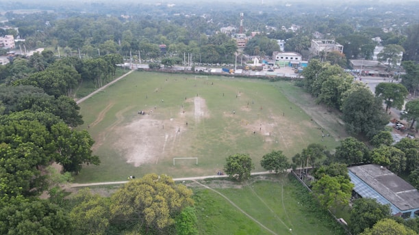An aerial view of a large open field surrounded by trees and urban structures. The field has visible patches of grass and dirt, with people scattered across, involved in various activities. A couple of goalposts suggest the area may be used for sports. Residential and commercial buildings are visible in the background, with a road running parallel to the field.