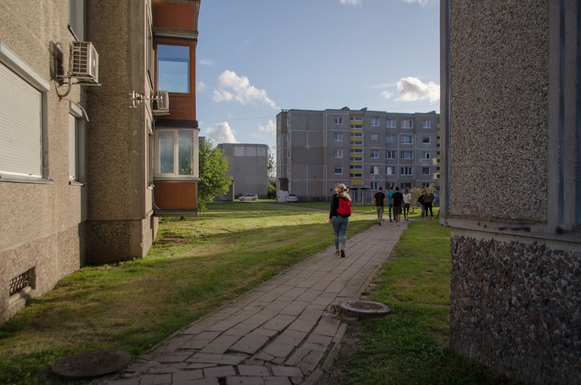 A residential area with two apartment buildings flanking a walkway. The path cuts through a grass lawn, and several people are walking along it. The sky is partly cloudy and the sun casts shadows on the buildings.