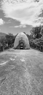A monochrome image capturing a dome-shaped structure in a natural setting. The structure features a mural of a seated figure on its front. Surrounding the building is a paved area leading up to it, with trees and foliage in the background under a partly cloudy sky.
