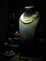 A dark display window showcases a variety of elegant jewelry pieces. The centerpiece is a black bust adorned with a double-strand pearl necklace featuring an elaborate floral pendant with green and red gemstones. Additional jewelry items, such as rings and brooches, are arranged on small pedestals to the left of the bust. The lighting highlights the luxurious and intricate details of the jewelry, creating a sophisticated and opulent atmosphere.