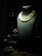 A dark display window showcases a variety of elegant jewelry pieces. The centerpiece is a black bust adorned with a double-strand pearl necklace featuring an elaborate floral pendant with green and red gemstones. Additional jewelry items, such as rings and brooches, are arranged on small pedestals to the left of the bust. The lighting highlights the luxurious and intricate details of the jewelry, creating a sophisticated and opulent atmosphere.