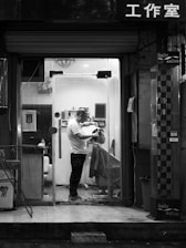 A barbershop scene captured at night in black and white. Inside a small, well-lit salon, a barber is cutting the hair of a seated client. The barber is standing and focused on the haircut, while the client is covered with a drape. Shelves with various hair products can be seen inside. The shopfront has a glass door, and there is Chinese lettering above it.