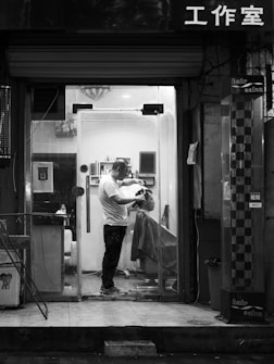 A barbershop scene captured at night in black and white. Inside a small, well-lit salon, a barber is cutting the hair of a seated client. The barber is standing and focused on the haircut, while the client is covered with a drape. Shelves with various hair products can be seen inside. The shopfront has a glass door, and there is Chinese lettering above it.