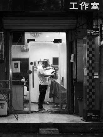 A barbershop scene captured at night in black and white. Inside a small, well-lit salon, a barber is cutting the hair of a seated client. The barber is standing and focused on the haircut, while the client is covered with a drape. Shelves with various hair products can be seen inside. The shopfront has a glass door, and there is Chinese lettering above it.