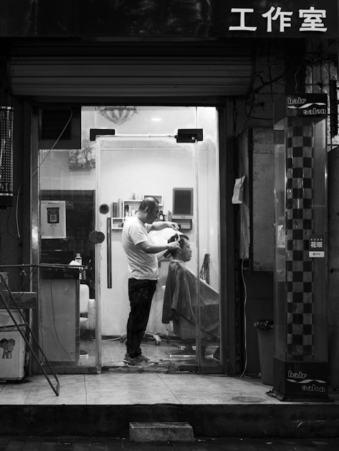A barbershop scene captured at night in black and white. Inside a small, well-lit salon, a barber is cutting the hair of a seated client. The barber is standing and focused on the haircut, while the client is covered with a drape. Shelves with various hair products can be seen inside. The shopfront has a glass door, and there is Chinese lettering above it.
