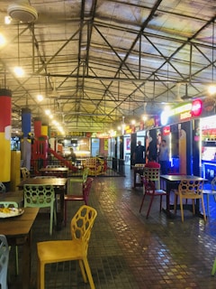A spacious indoor food court with a high ceiling and metal beams, featuring brightly colored chairs and tables. Various food stalls with illuminated signs line the sides, and a children's play area with a slide is visible. The floor is made of small tiles, and the space is lit by overhead lights.