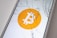 A smartphone screen displaying the Bitcoin logo is cracked, with multiple fractures radiating from the center of the screen.