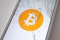 A smartphone screen displaying the Bitcoin logo is cracked, with multiple fractures radiating from the center of the screen.