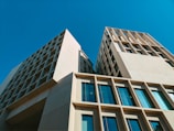 Modern architectural buildings with geometric designs under a clear blue sky. The structures feature extensive use of glass windows and beige stone façades, emphasizing clean lines and symmetry.