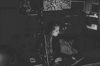 A black and white photo features a woman seated on a couch, focused on a laptop. The room appears dimly lit with a microphone stand in the foreground. In the background, there are stacked firewood and computer monitors on a shelf, creating a cozy and tech-influenced workspace.