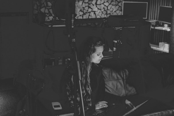 A black and white photo features a woman seated on a couch, focused on a laptop. The room appears dimly lit with a microphone stand in the foreground. In the background, there are stacked firewood and computer monitors on a shelf, creating a cozy and tech-influenced workspace.