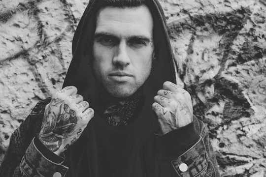 A black and white portrait of a person with tattoos, wearing a hooded jacket. The person is holding the hood up with both hands. The background is textured, possibly a wall with graffiti.