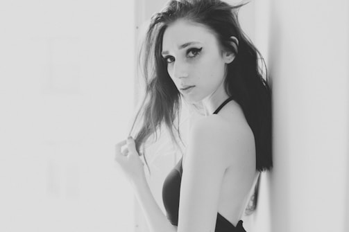 A monochrome photograph featuring a woman with dark hair and striking eye makeup, standing in a minimalistic setting. She gazes directly at the camera with a subtle expression, creating an intimate and artistic atmosphere.