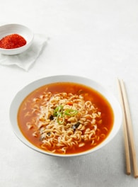A bowl of ramen noodles is served in a white bowl, filled with bright red, spicy-looking broth and topped with sliced green chilies and mushrooms. Wooden chopsticks are placed to the right of the bowl on a light surface. In the background, a small white dish containing red chili flakes is placed on a white napkin.