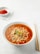 A bowl of ramen noodles is served in a white bowl, filled with bright red, spicy-looking broth and topped with sliced green chilies and mushrooms. Wooden chopsticks are placed to the right of the bowl on a light surface. In the background, a small white dish containing red chili flakes is placed on a white napkin.