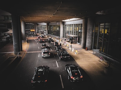 A line of cars is waiting in front of a modern building with large glass windows and tall pillars. Several people are near the entrance, some carrying luggage. The scene appears to be a drop-off or pick-up area, possibly at an airport.