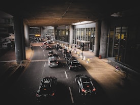 A line of cars is waiting in front of a modern building with large glass windows and tall pillars. Several people are near the entrance, some carrying luggage. The scene appears to be a drop-off or pick-up area, possibly at an airport.