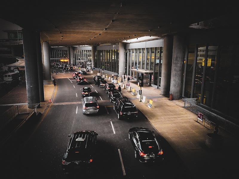 A line of cars is waiting in front of a modern building with large glass windows and tall pillars. Several people are near the entrance, some carrying luggage. The scene appears to be a drop-off or pick-up area, possibly at an airport.