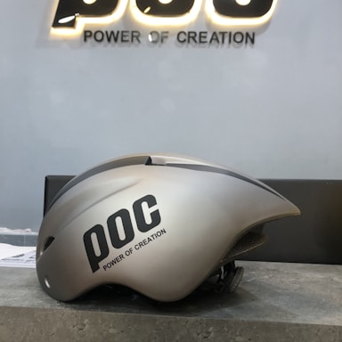 A sleek, metallic gray helmet with a streamlined design is placed on a textured surface. The helmet has the letters 'POC' and the phrase 'Power of Creation' printed on its side in black. Behind the helmet, the same 'POC Power of Creation' text is illuminated on a gray wall.