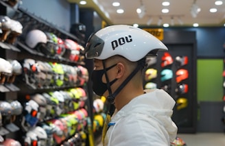 A person wearing a white helmet is standing in a store that sells various helmets. The person is facing sideways and is wearing a black face mask. Shelves in the background display a wide variety of helmets in different colors and styles, creating a vibrant and busy atmosphere.