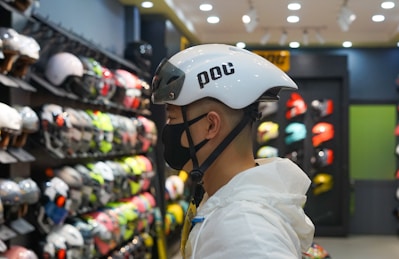 A person wearing a white helmet is standing in a store that sells various helmets. The person is facing sideways and is wearing a black face mask. Shelves in the background display a wide variety of helmets in different colors and styles, creating a vibrant and busy atmosphere.