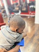 A young child wearing a gray shirt is being held closely. The background features a barber shop setting with blurred figures and red accents.