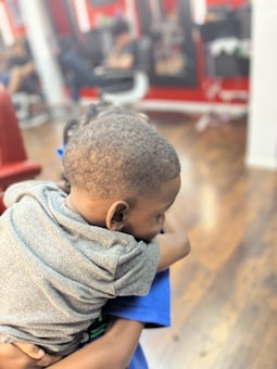 A young child wearing a gray shirt is being held closely. The background features a barber shop setting with blurred figures and red accents.