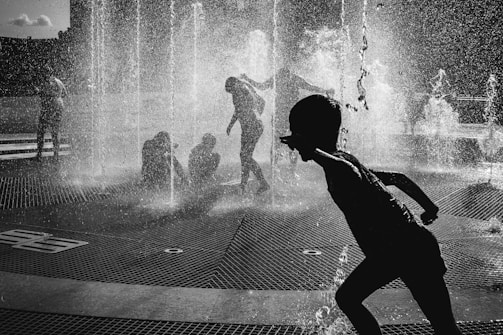 Children are playing joyfully in an urban water fountain, with streams of water creating a dynamic and lively atmosphere. The silhouettes of the children are captured in various playful poses against the backdrop of sprinkling water, conveying a sense of movement and excitement.
